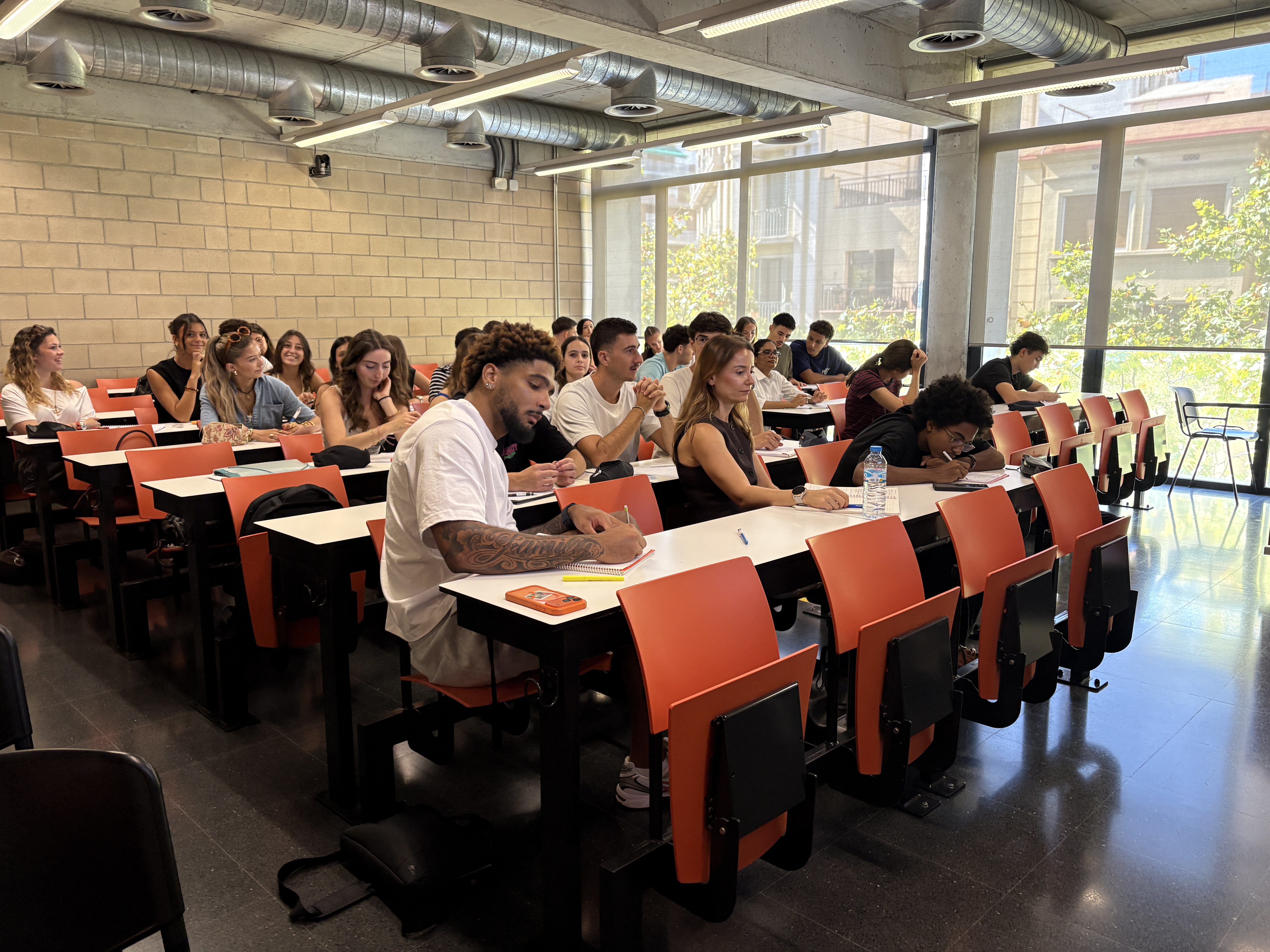 L'alumnat a l'aula durant els cursos de català. La majoria d'estudiants estan concentrats, però hi ha una noia al fons que està mirant a càmera amb el somriure més gran que te pots imaginar