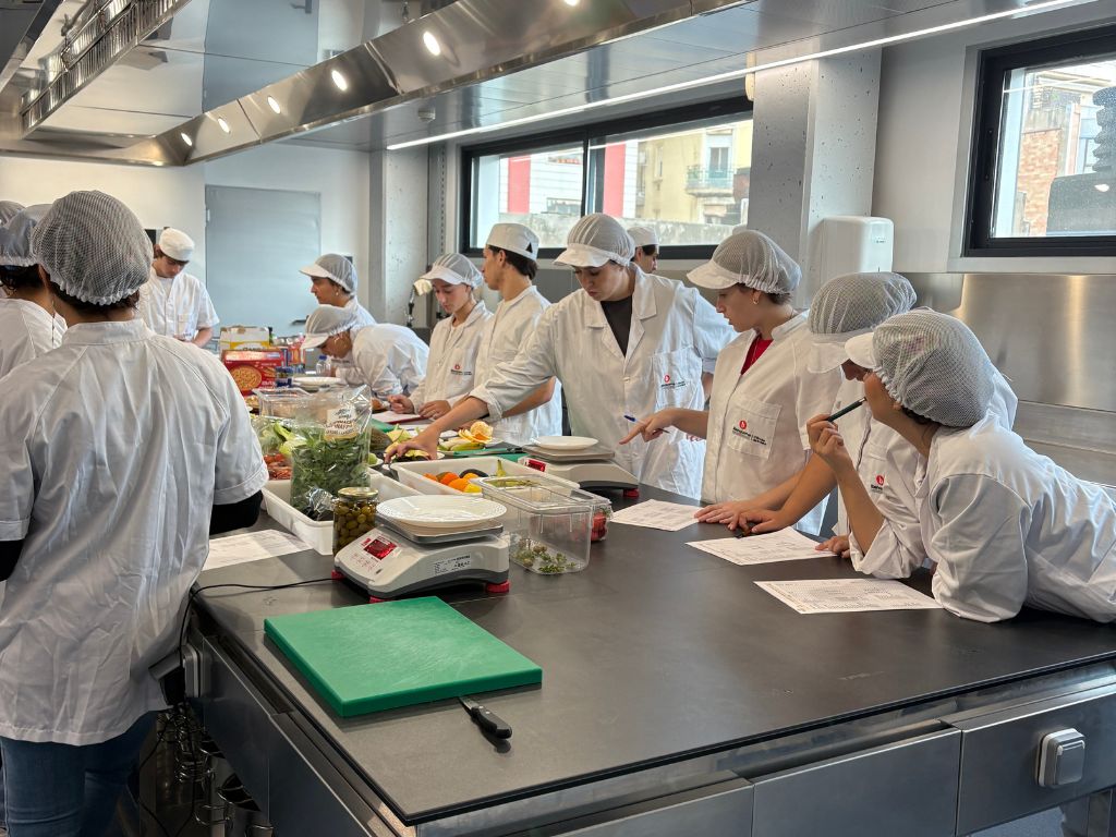 L'alumnat està a la cuina treballant amb aliments, que estan tots situats a la meitat de la taula. L'alumnat que no està d'esquenes, està parlant entre ells.