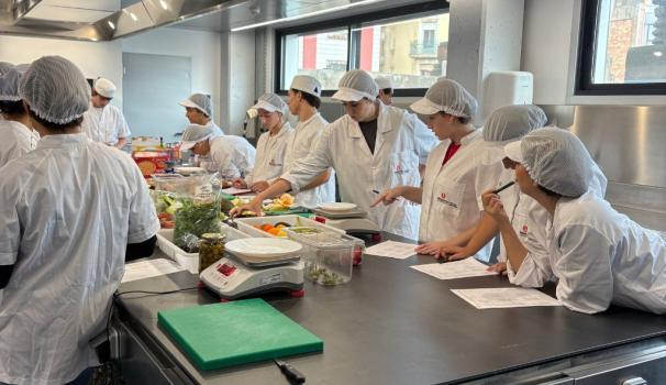 L'alumnat està a la cuina treballant amb aliments, que estan tots situats a la meitat de la taula. L'alumnat que no està d'esquenes, està parlant entre ells.